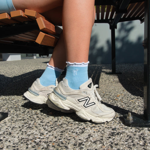 Person wearing light blue socks and white sneakers with a visible 'N' logo, sitting on a bench.