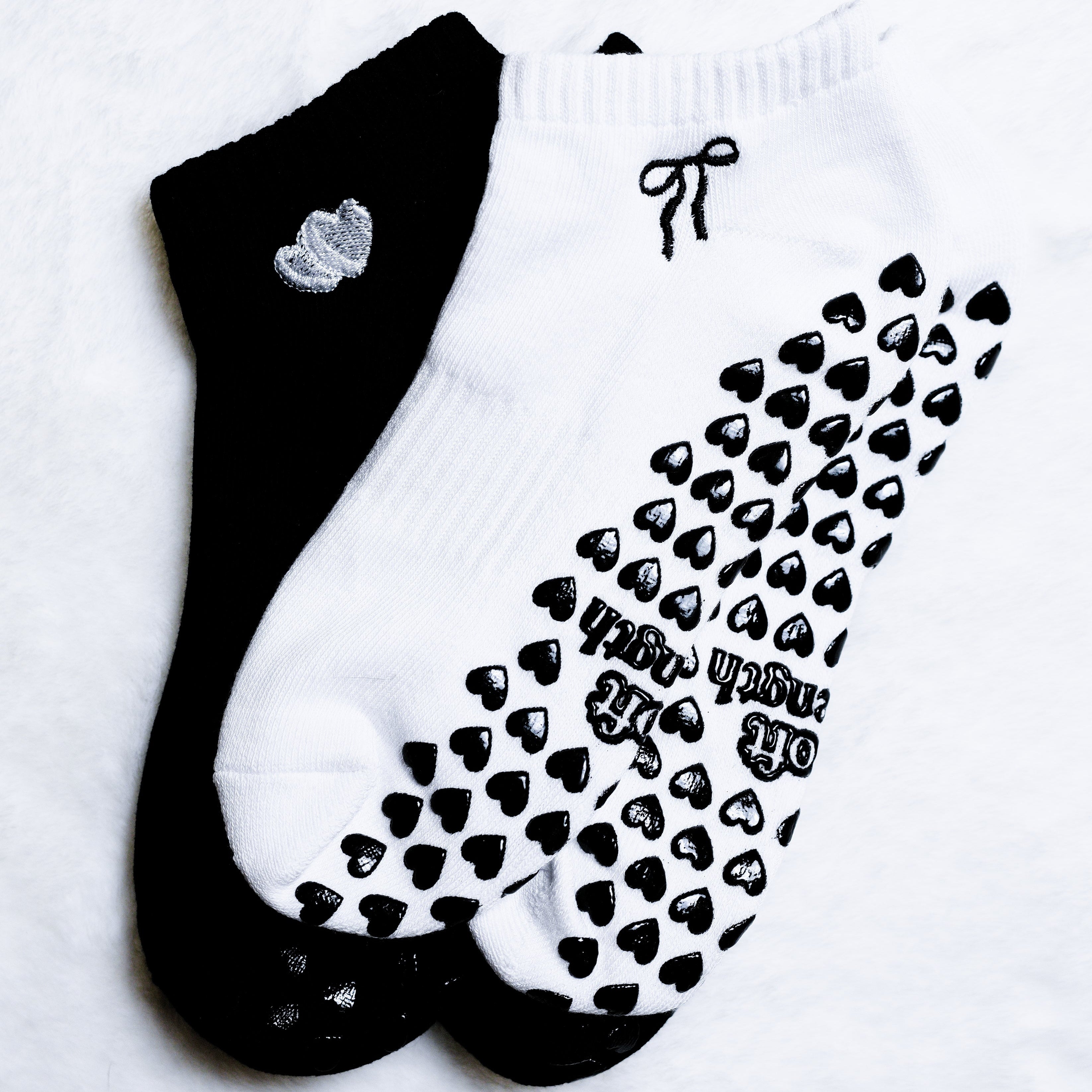 Two ankle socks laid flat on a white textured background: one black sock with a small silver heart detail, and one white sock with black heart grip pads and the words 'HAPPY TRASH DAY' printed on the sole.