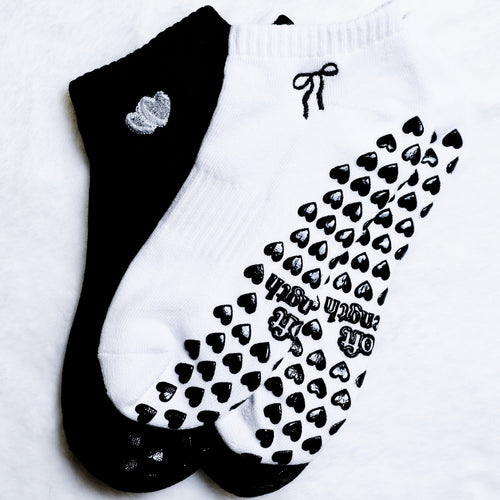 Two ankle socks laid flat on a white textured background: one black sock with a small silver heart detail, and one white sock with black heart grip pads and the words 'HAPPY TRASH DAY' printed on the sole.