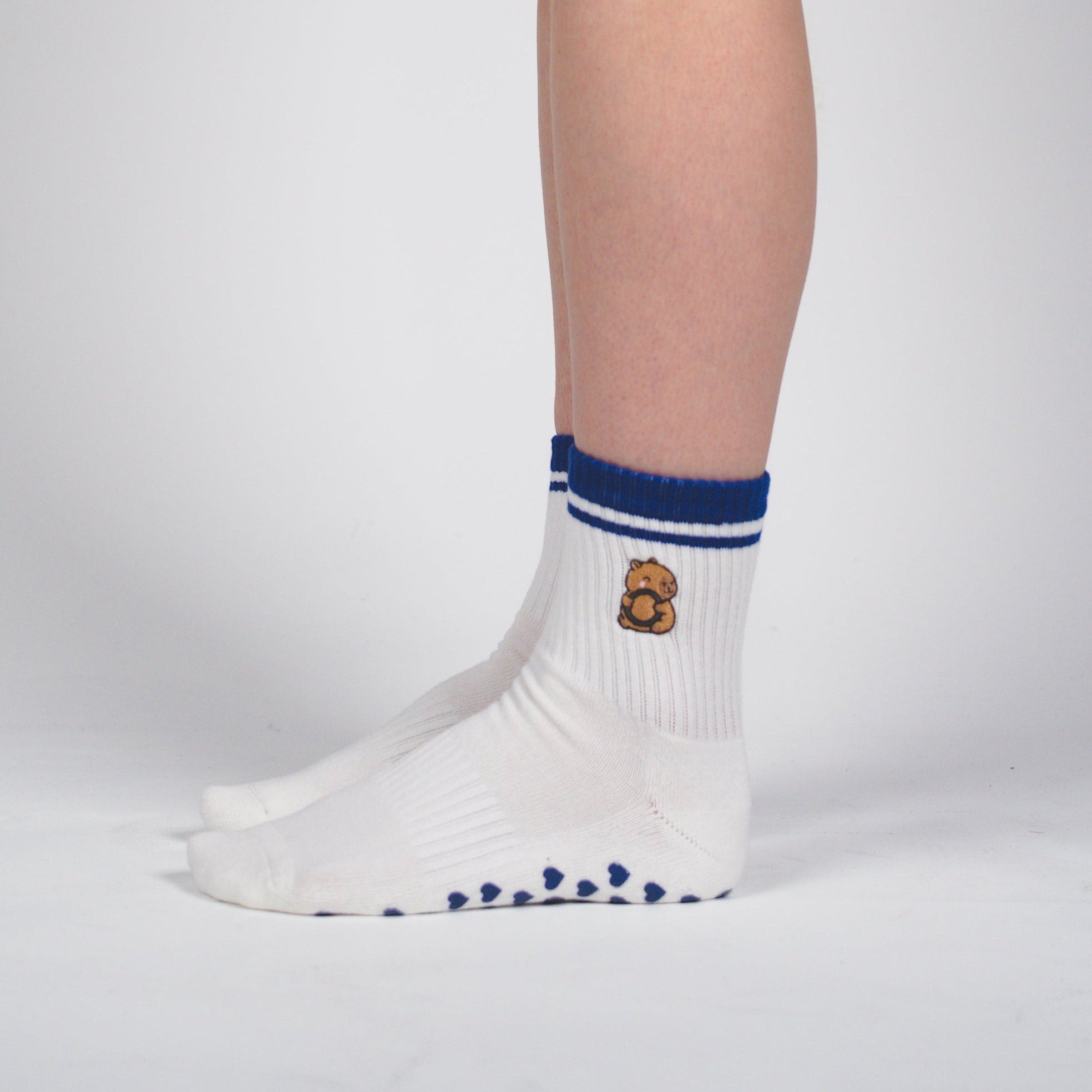 A side profile of a person's leg wearing a white athletic sock with blue trim and a small embroidered logo, paired with black shorts, against a light gray studio background.