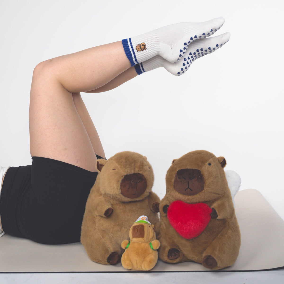 A person lying on a yoga mat with one leg raised, wearing white socks with blue grip dots. In front of them are two brown capybara plush toys, one holding a red heart and one holding a small green item, against a plain light gray studio background.