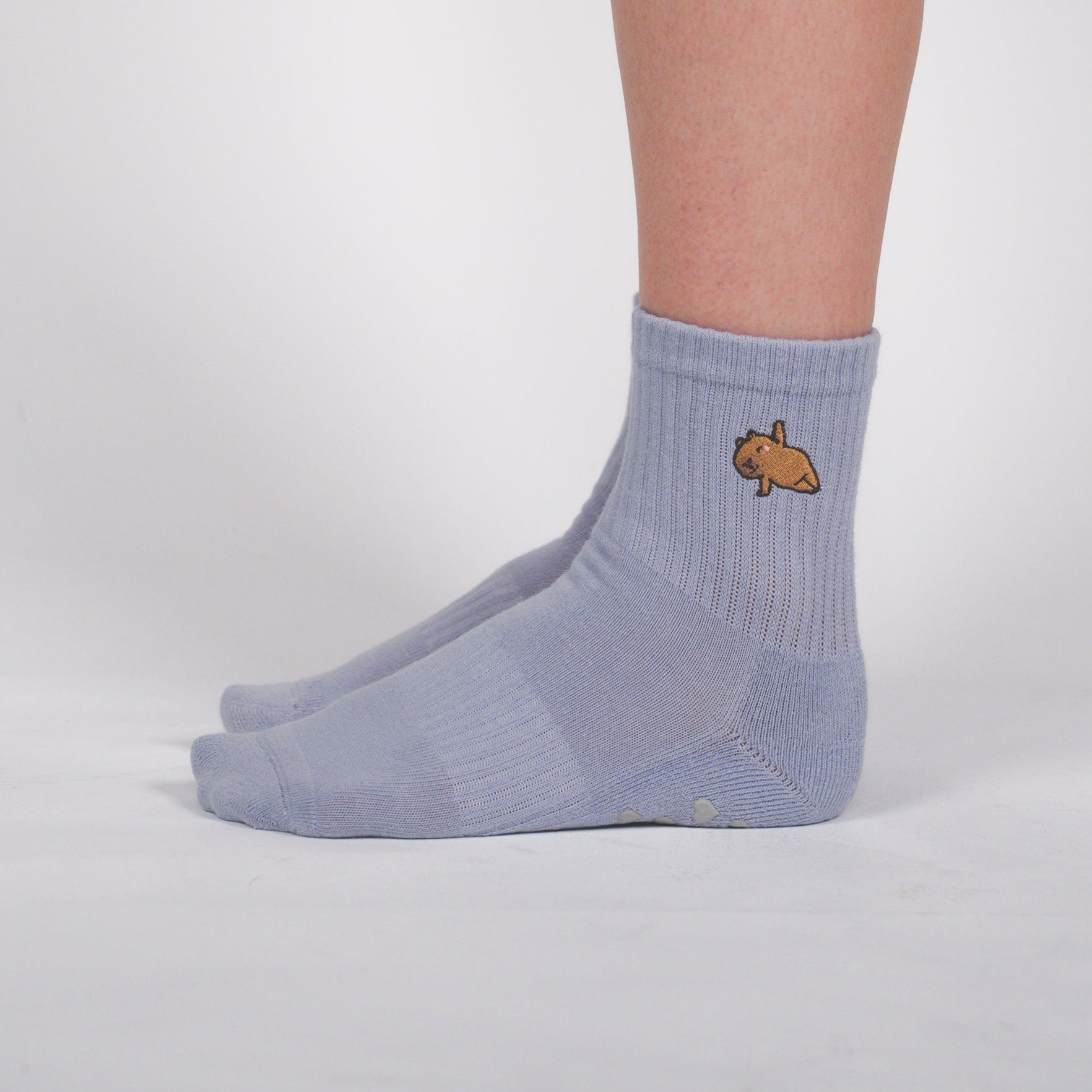 A person's lower leg and foot wearing a light gray ribbed ankle sock with a small embroidered fish logo on the side, standing against a light gray studio background.