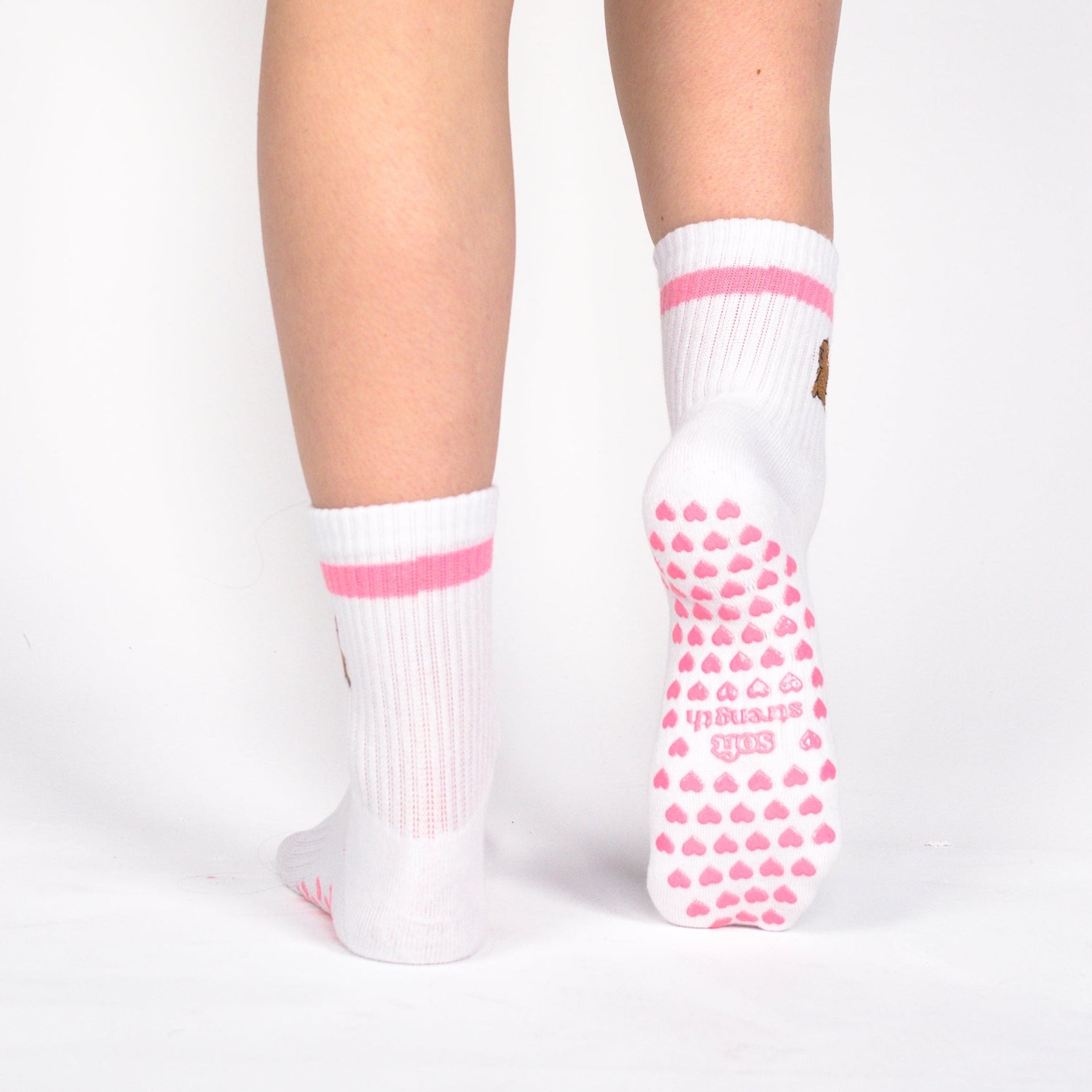 Rear view of a person wearing black athletic shorts, white crew socks with pink stripes, and white sneakers with pink grip dots, standing against a light gray studio background.