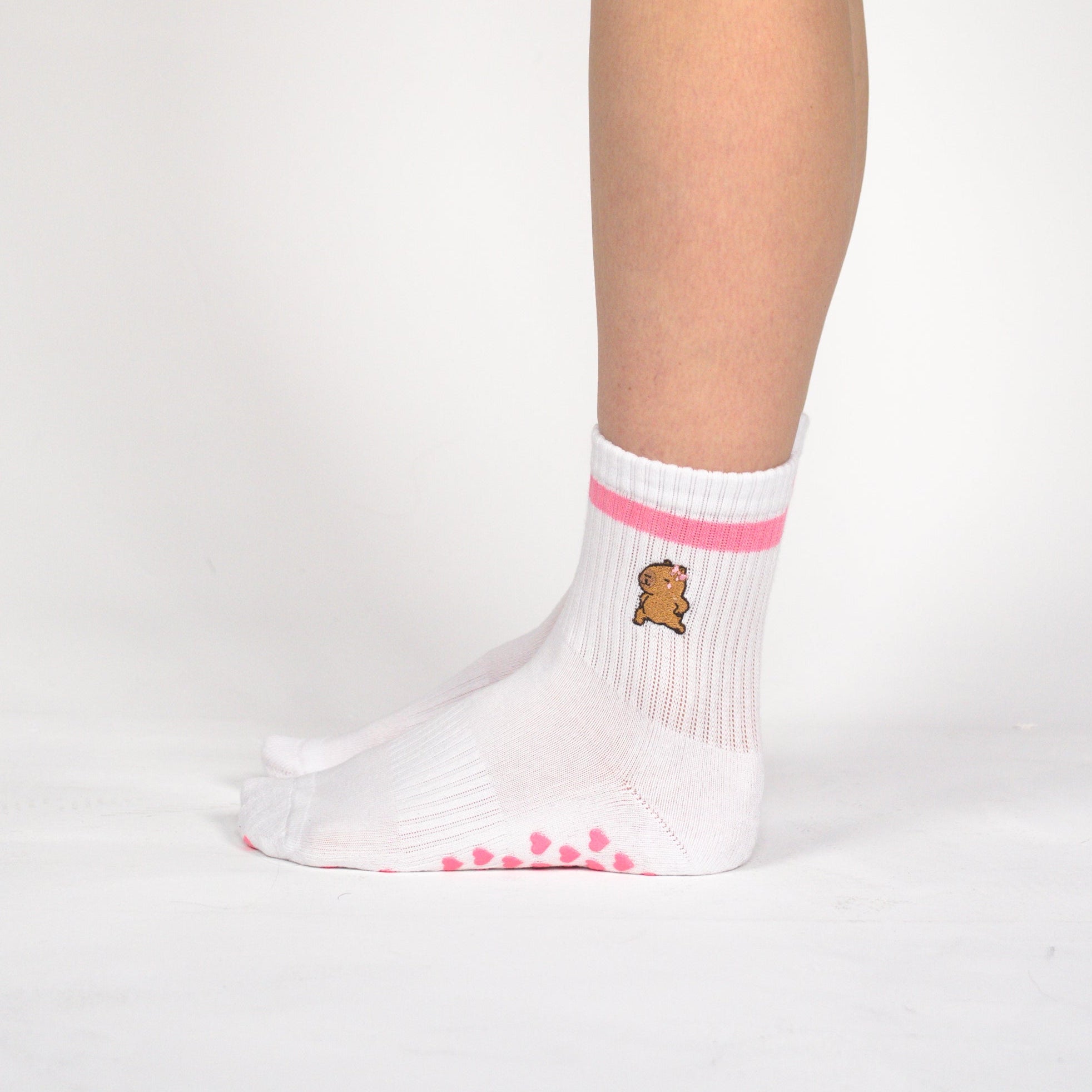 A person's lower leg and foot wearing a white grip sock with pink stripes and a small teddy bear graphic, paired with black athletic shorts against a light gray studio background.
