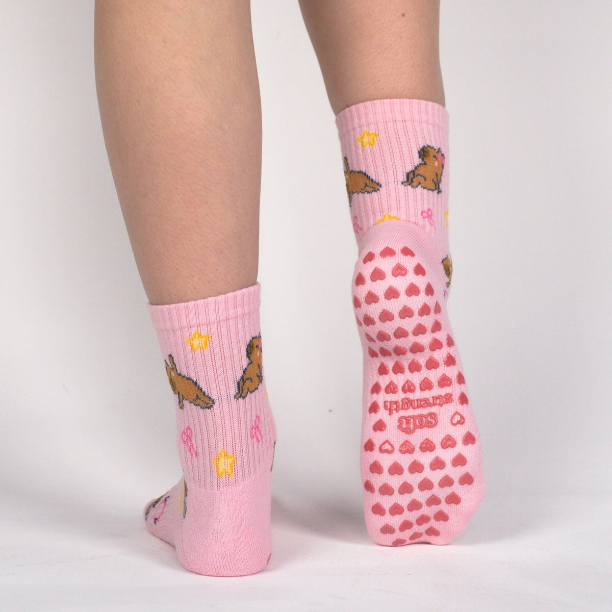 Pink biker shorts worn by a model, shown from the back against a light gray studio background. The model is wearing pink socks with a brown and yellow pattern on one sock and a pink grip sole with red dots on the other.