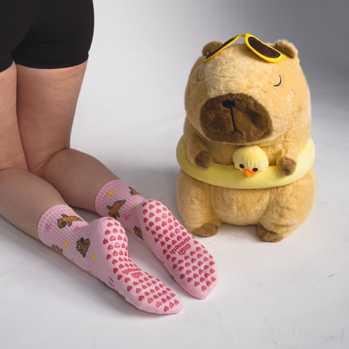 Pink grip socks with brown bear prints worn by a model, shown next to a plush toy in a studio setting.