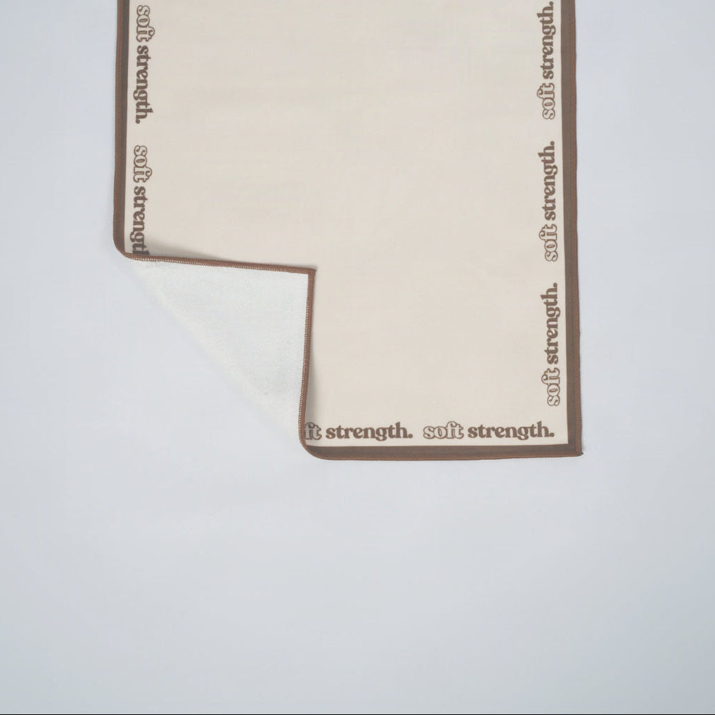 A cream-colored fabric mat with a brown border printed with the words 'soft strength.' along the edges, shown on a plain white background with one corner folded over.